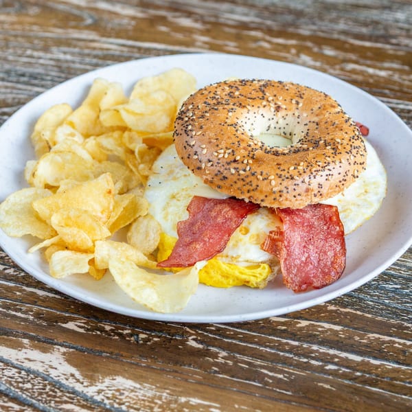Everything bagel sandwich with fried eggs, bacon, and a side of potato chips on a white plate.