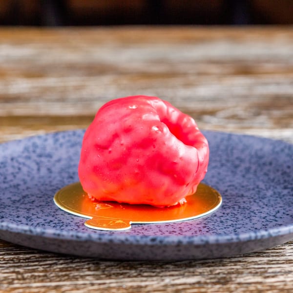 A vibrant red glazed dessert sits on a gold coaster atop a speckled blue plate.