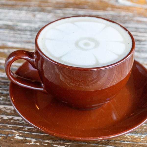 A brown cup and saucer with a latte featuring a flower design in the foam on a wooden table.