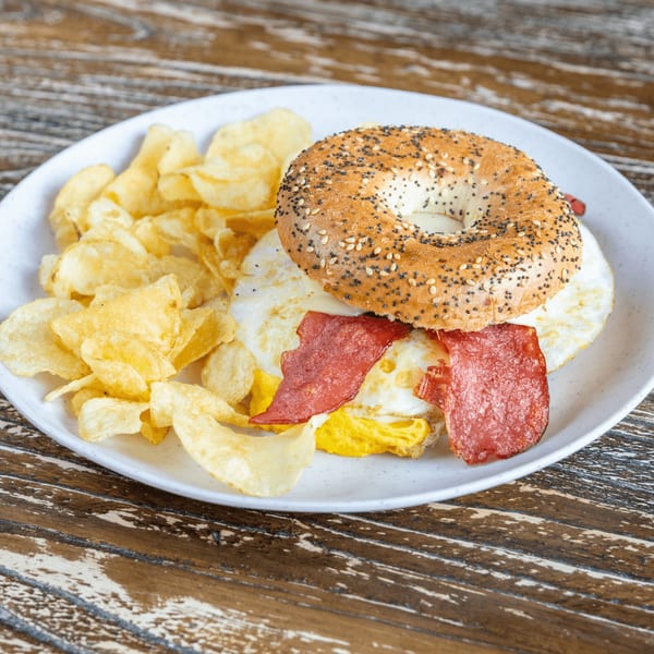 Everything bagel breakfast sandwich with fried eggs, bacon, and a side of potato chips.
