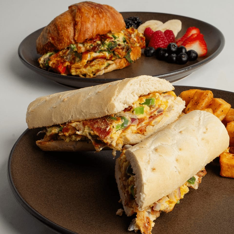 Two breakfast plates: a croissant sandwich with fruit, and a baguette sandwich with home fries.