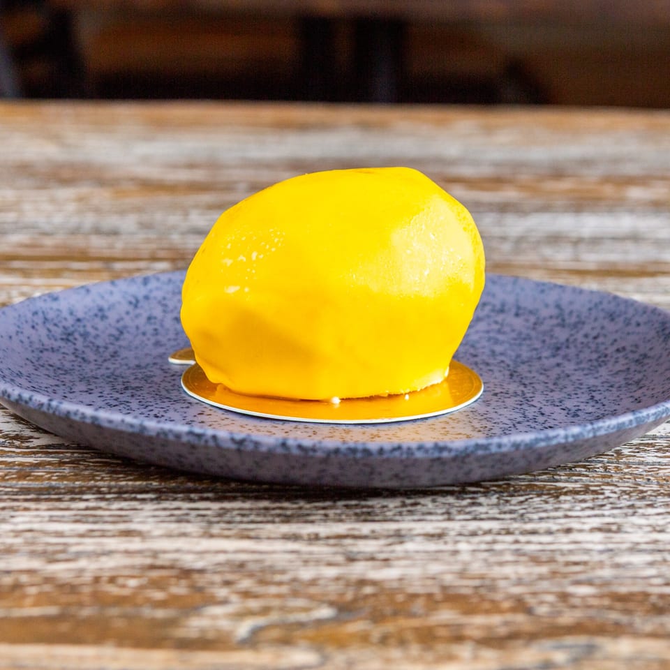 A bright yellow lemon-shaped dessert with a glossy glaze sits on a dark blue plate.