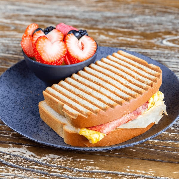 Grilled sandwich with egg, bacon, and cheese, served with a side of fresh strawberries and blueberries.