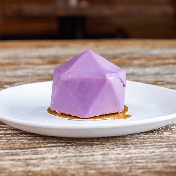 A purple geometric dessert with glaze on a white plate, against a rustic wooden background.