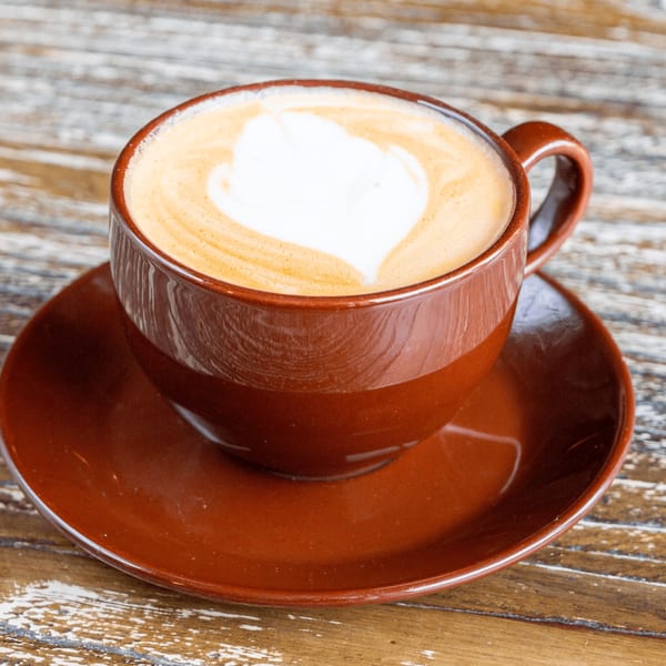 A brown ceramic cup and saucer hold a latte with a heart-shaped foam design on a wooden table.