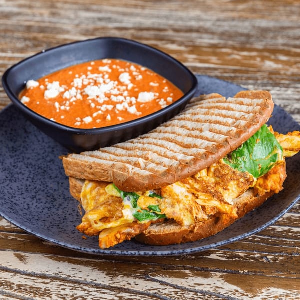 A grilled sandwich with scrambled eggs and greens, served with a bowl of tomato soup topped with feta cheese.