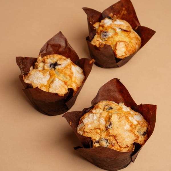 Three blueberry muffins with brown wrappers and a sugar topping on a tan surface.