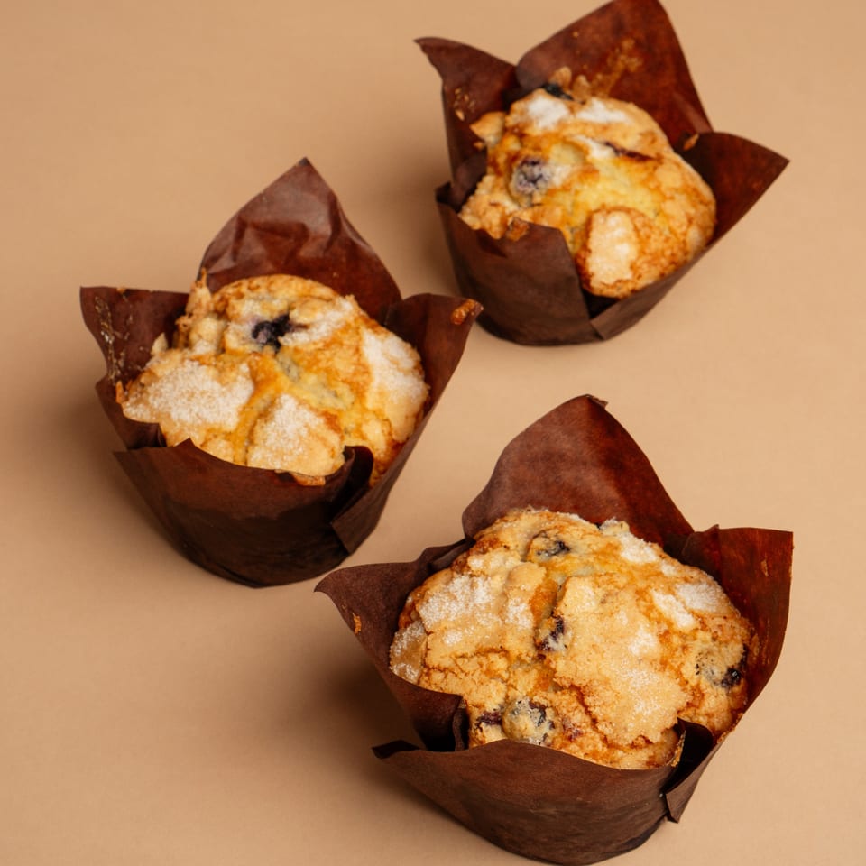 Three blueberry muffins with brown wrappers and a sugar topping on a tan surface.