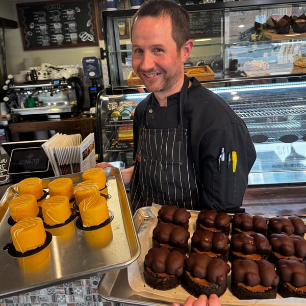 A smiling Rendezvous International Cafe employee holds trays of yellow petit fours and chocolate desserts.