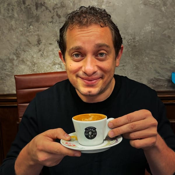 Man smiling and holding a small white cup of coffee with a saucer at Rendezvous International Cafe.