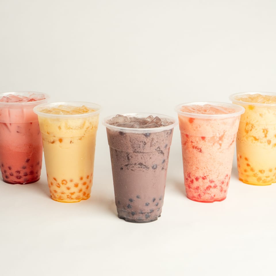 Five colorful bubble tea drinks with tapioca pearls and ice in clear plastic cups.