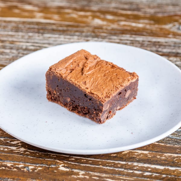 A rich, dark chocolate brownie with a crinkled top sits on a white plate on a wooden table.