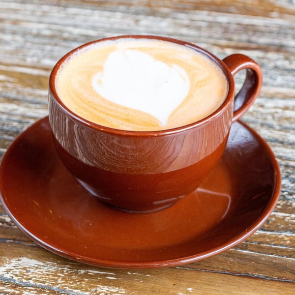 A brown cup of latte with heart-shaped foam art sits on a matching saucer on a wooden table.