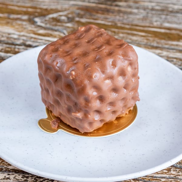 A cube-shaped chocolate dessert with a textured, nutty coating sits on a white plate.