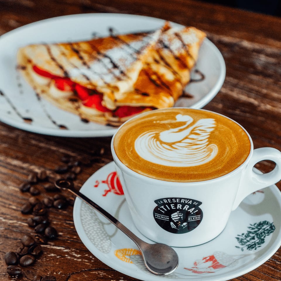 A latte with swan art and a strawberry crepe on a wooden table at Rendezvous International Cafe.